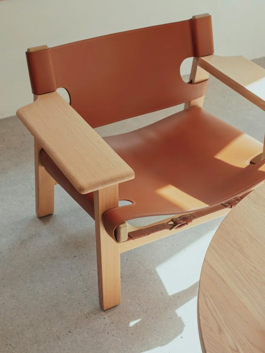 Stol med lyst trestativ og karamellbrunt skinnsete og rygg, med synlige spenner. Et rundt trebord er delvis synlig.