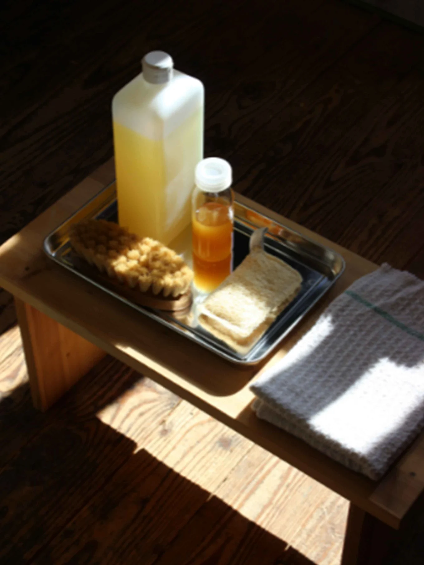 Badeartikler på et brett: Flaske gul væske, flaske oransje væske, badebørste, loofah. Hvitt håndkle på trebord.