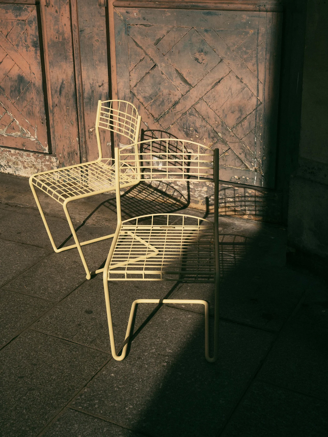To lysegule metallstoler på fliser foran en slitt tredør med geometrisk mønster, med harde skygger.