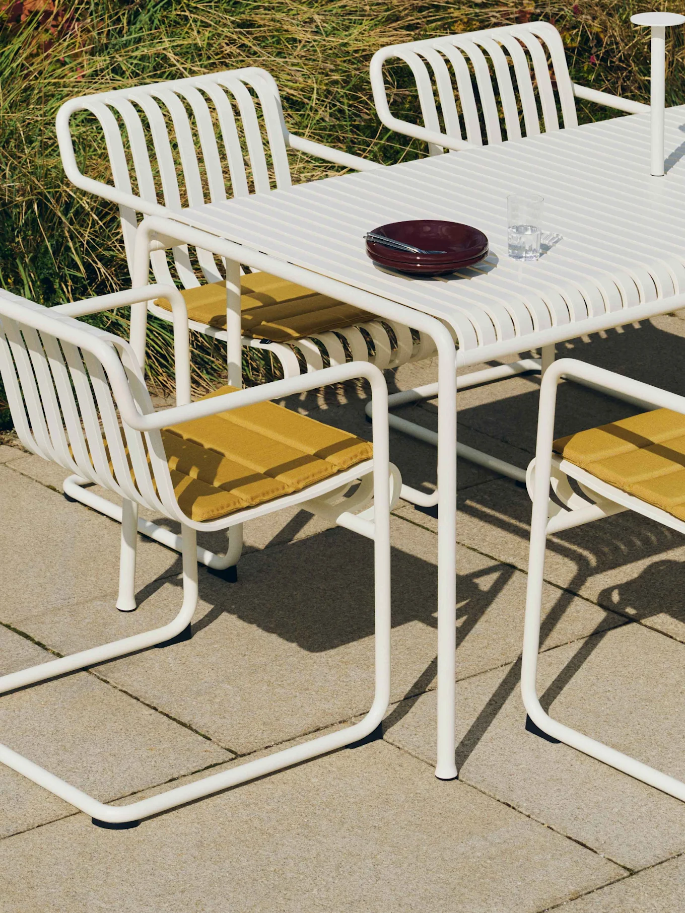 Hvit lamellbord med matchende stoler, gule puter og servise på et utendørs gårdsrom.